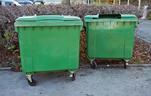 Two large green plastic wheeled rubbish bins are positioned on a paved surface, likely a driveway or street in an urban or suburban area. The bins have hinged lids and are made of durable, slightly textured plastic with a matte finish. They are placed side by side, with the left bin slightly larger than the right, and both are positioned in front of a background of leafless shrubbery and a low metal fence. The environment appears clean and outdoors, with some fallen leaves scattered on the ground. The bins are typical for commercial waste collection purposes, consistent with rubbish removal services offered by companies such as Commercial Waste Amersham. The scene suggests routine waste disposal activity, with the bins ready for collection or emptying, emphasizing the focus on waste management solutions in the local area near Amersham postcode or town.