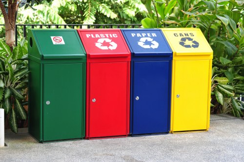 A row of four recycling bins placed on a paved outdoor area with lush green foliage in the background. The bins are arranged side by side, each with a distinct colour and label indicating the type of waste: green for general waste, red for plastic, blue for paper, and yellow for cans. Each bin features a round silver lock in the lower central part of the front panel, and the lids are slightly angled to facilitate waste disposal. The surface of the bins appears to be made of metal or durable plastic with a smooth finish, and they are positioned on a concrete or asphalt surface, typical for waste collection points. The environment suggests a community or commercial area in Amersham, with the background filled with leaves and branches, emphasizing an outdoor setting aligned with rubbish removal services offered by Commercial Waste Amersham.